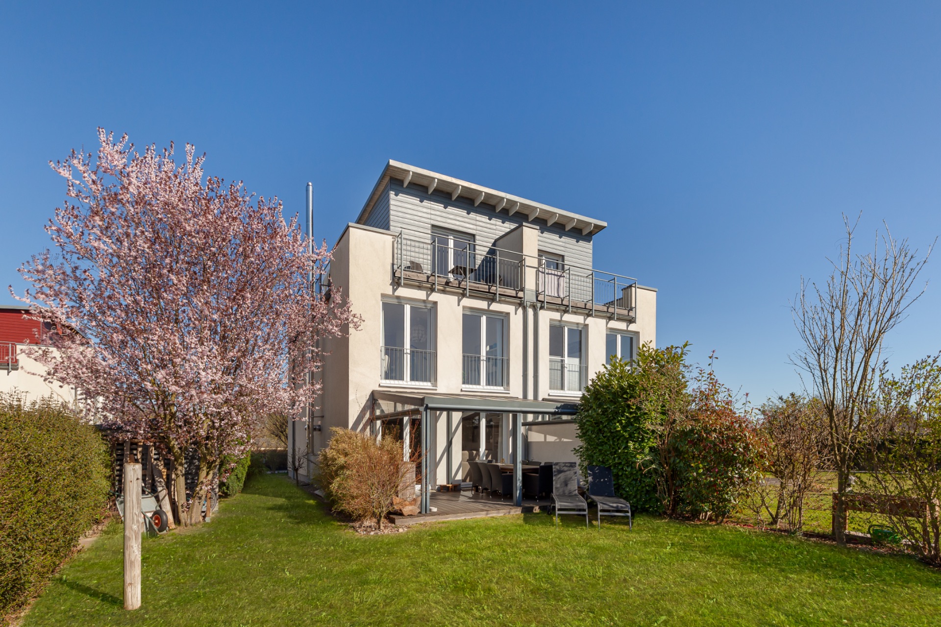 Modernes dreistöckiges Wohnhaus mit Balkon, Garten und blühendem Baum im Vordergrund.