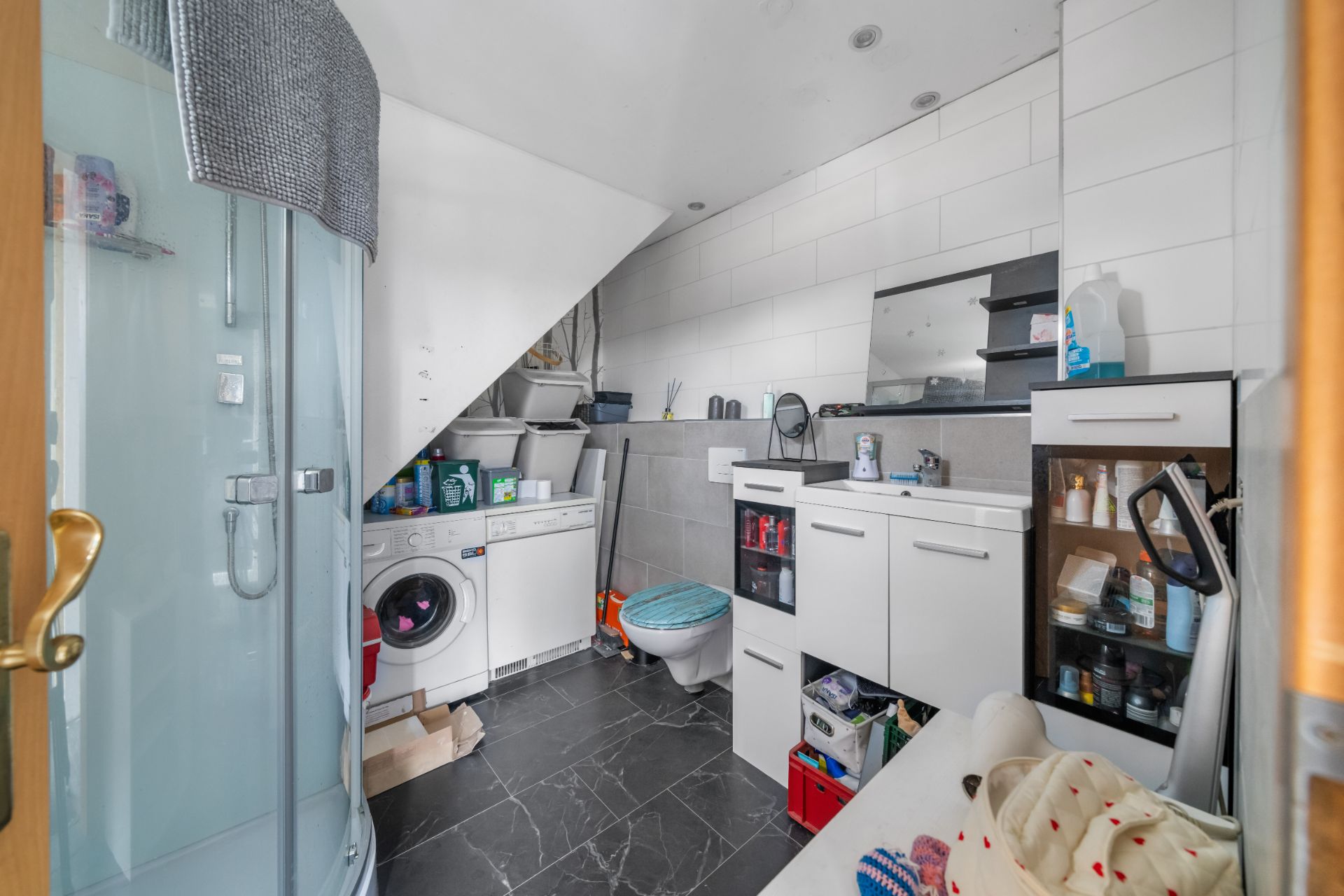 Modernes Badezimmer mit Dusche, Waschmaschine, WC und weißen Schränken unter einer Dachschräge.
