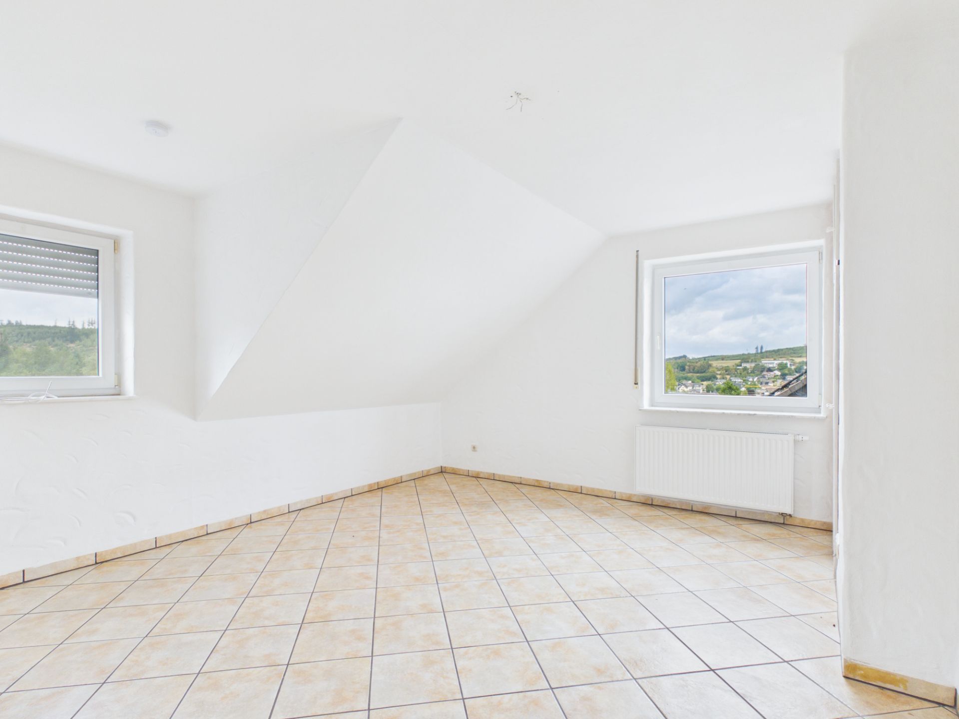 Heller Raum mit Dachschräge, Fliesenboden und zwei Fenstern mit Ausblick.