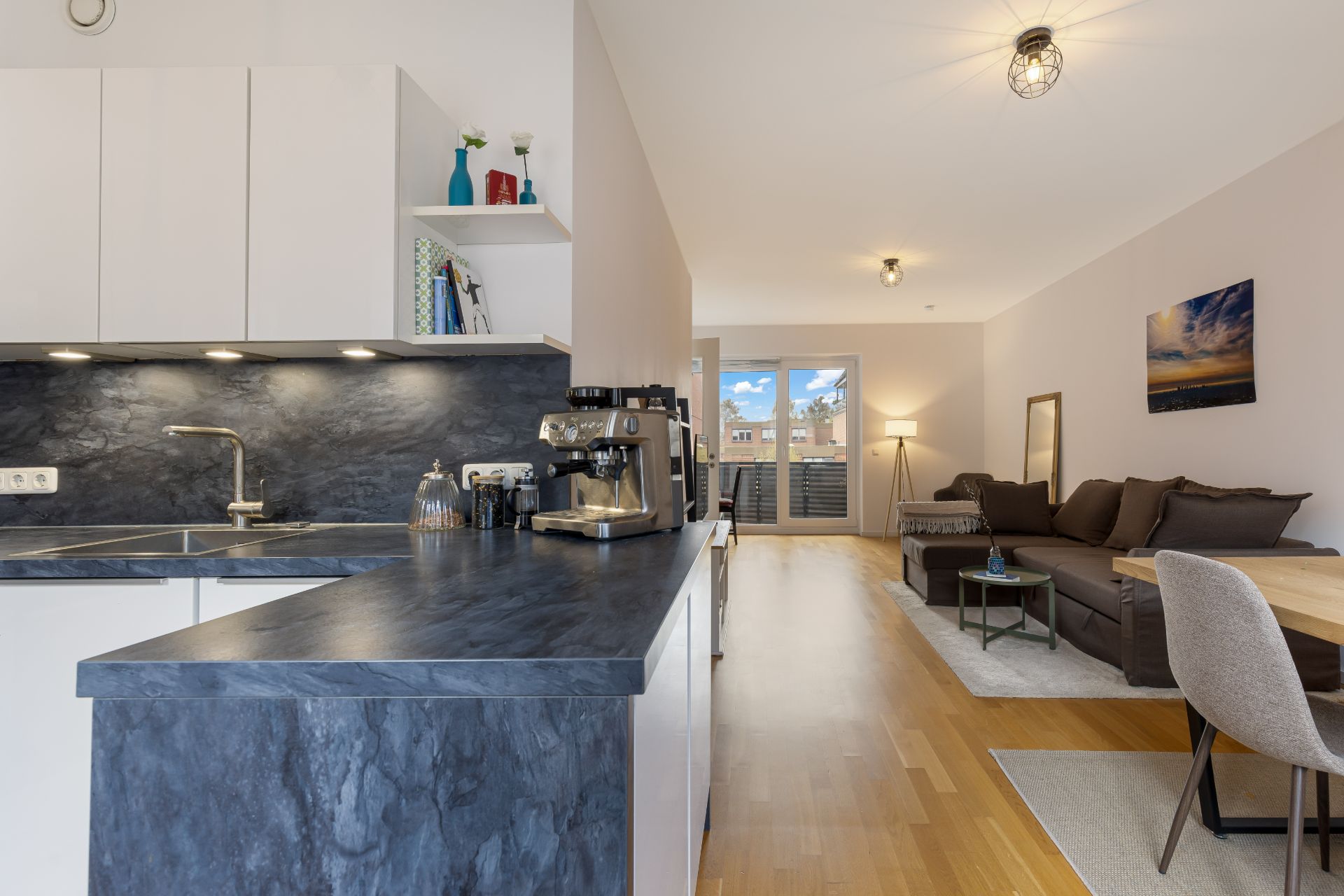 Moderne Küche mit dunkler Arbeitsplatte und offenem Wohnzimmer mit Sofa und Esstisch.