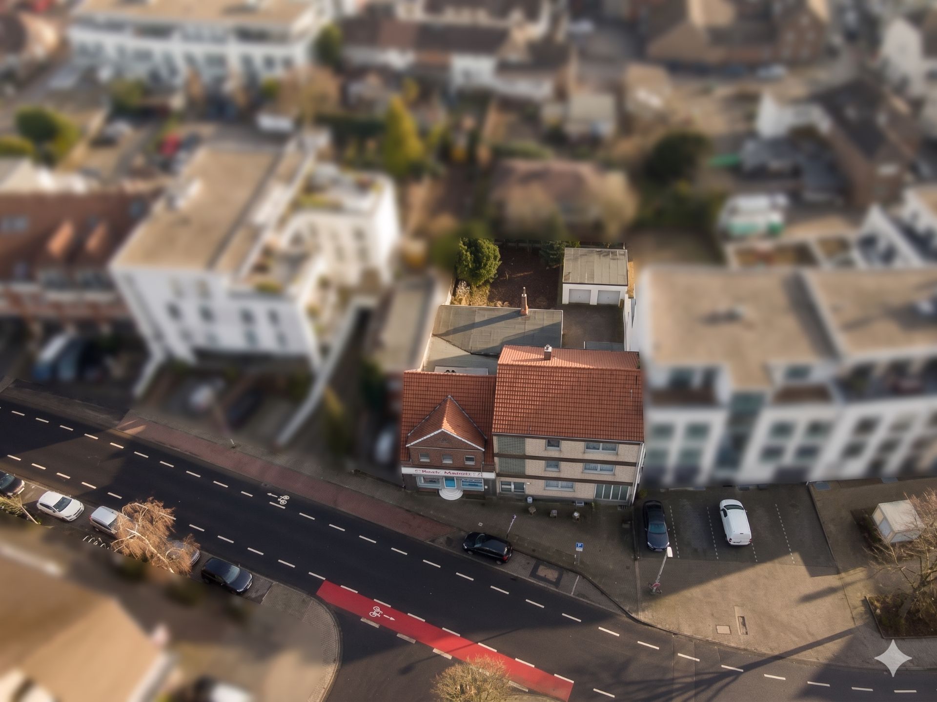 Luftaufnahme eines Wohnhauses mit rotem Dach an einer Straße, daneben Parkplatz und Nachbargebäude.