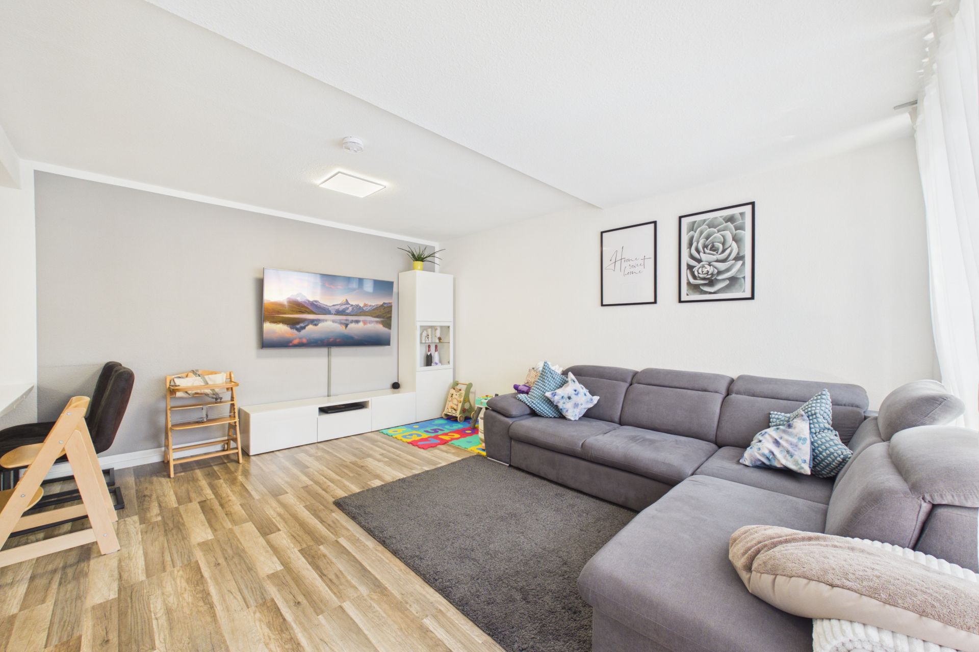 Helles Wohnzimmer mit grauem Ecksofa, Fernseher, Spielteppich und Holzboden.