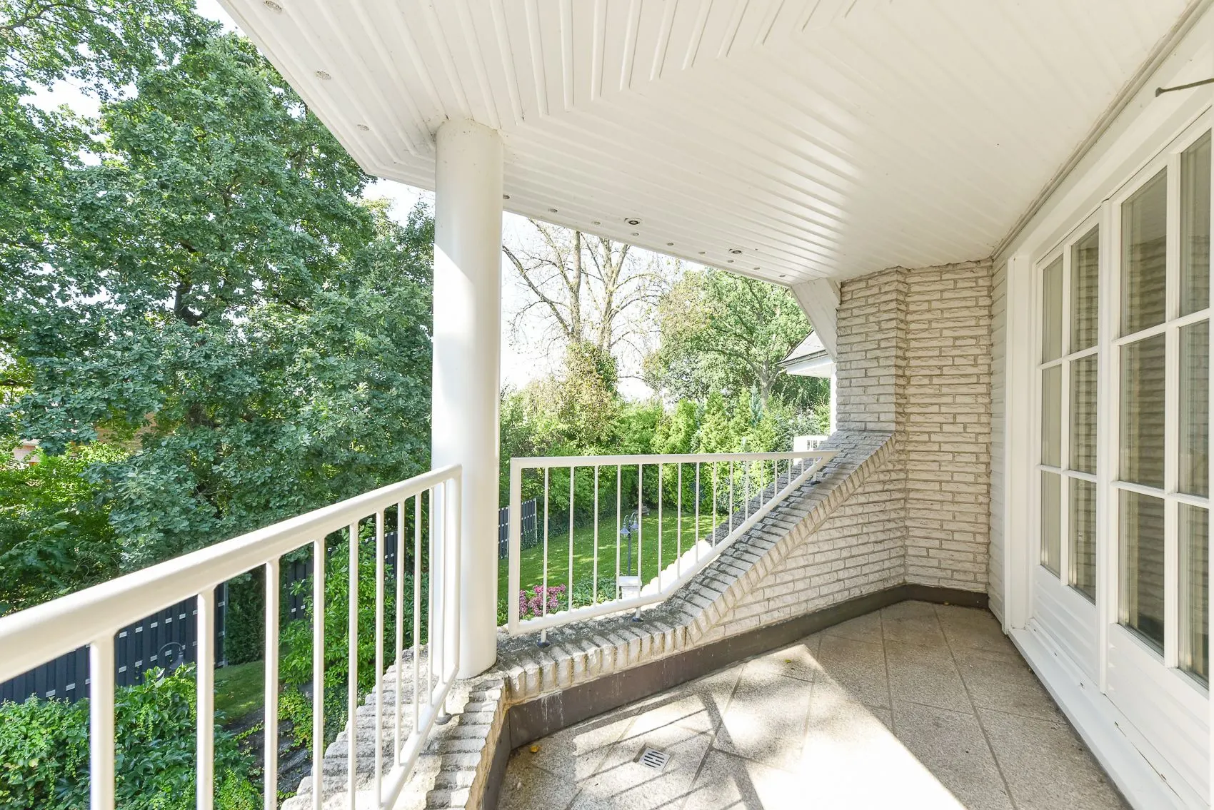 Überdachter Balkon mit Geländer, Fliesenboden und Blick auf grünen Garten mit Bäumen.