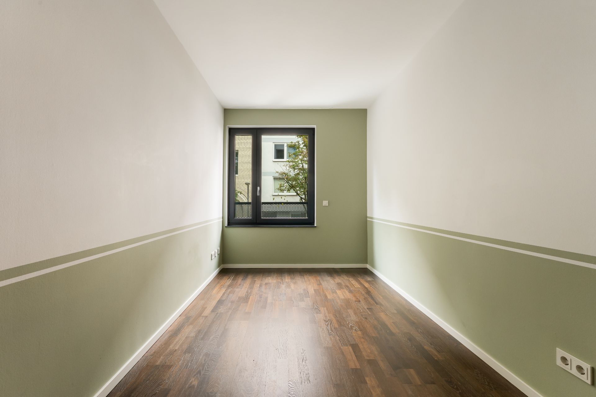 Leerer, schmaler Raum mit grün-weißen Wänden, Holzfußboden und Fenster mit Blick nach draußen.