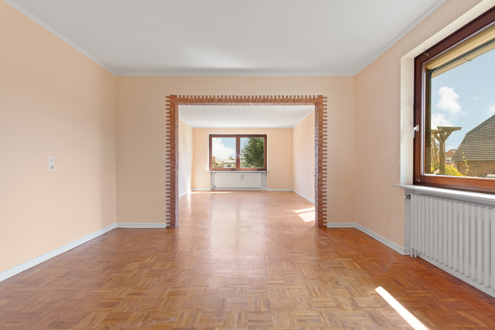 Leerer Raum mit Parkettboden, großen Fenstern und einer offenen Verbindung zum nächsten Zimmer.