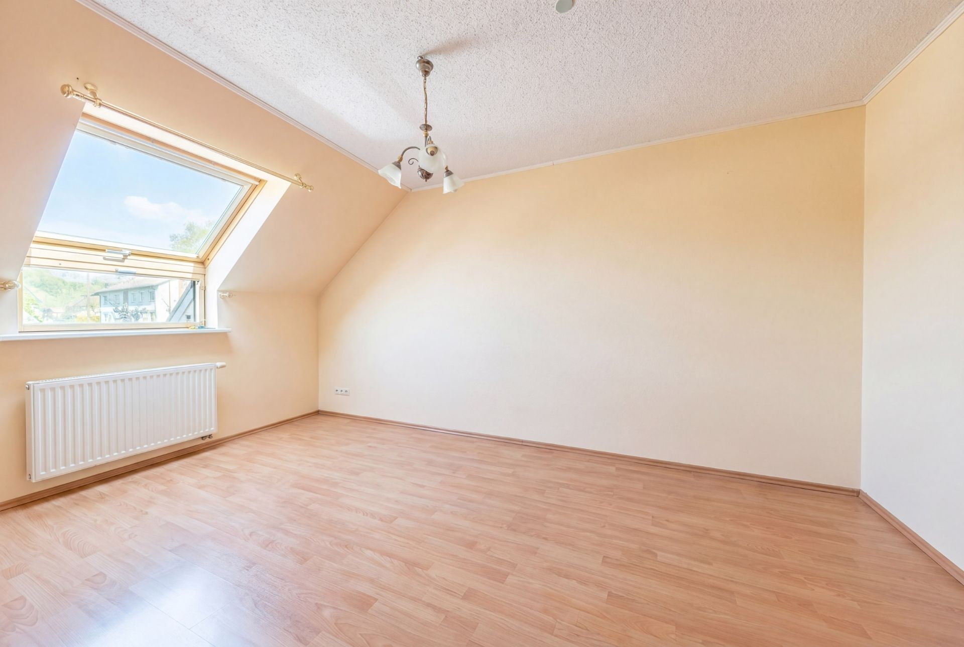 Leeres Zimmer mit schräger Decke, großem Fenster, Heizkörper und Holzfußboden.