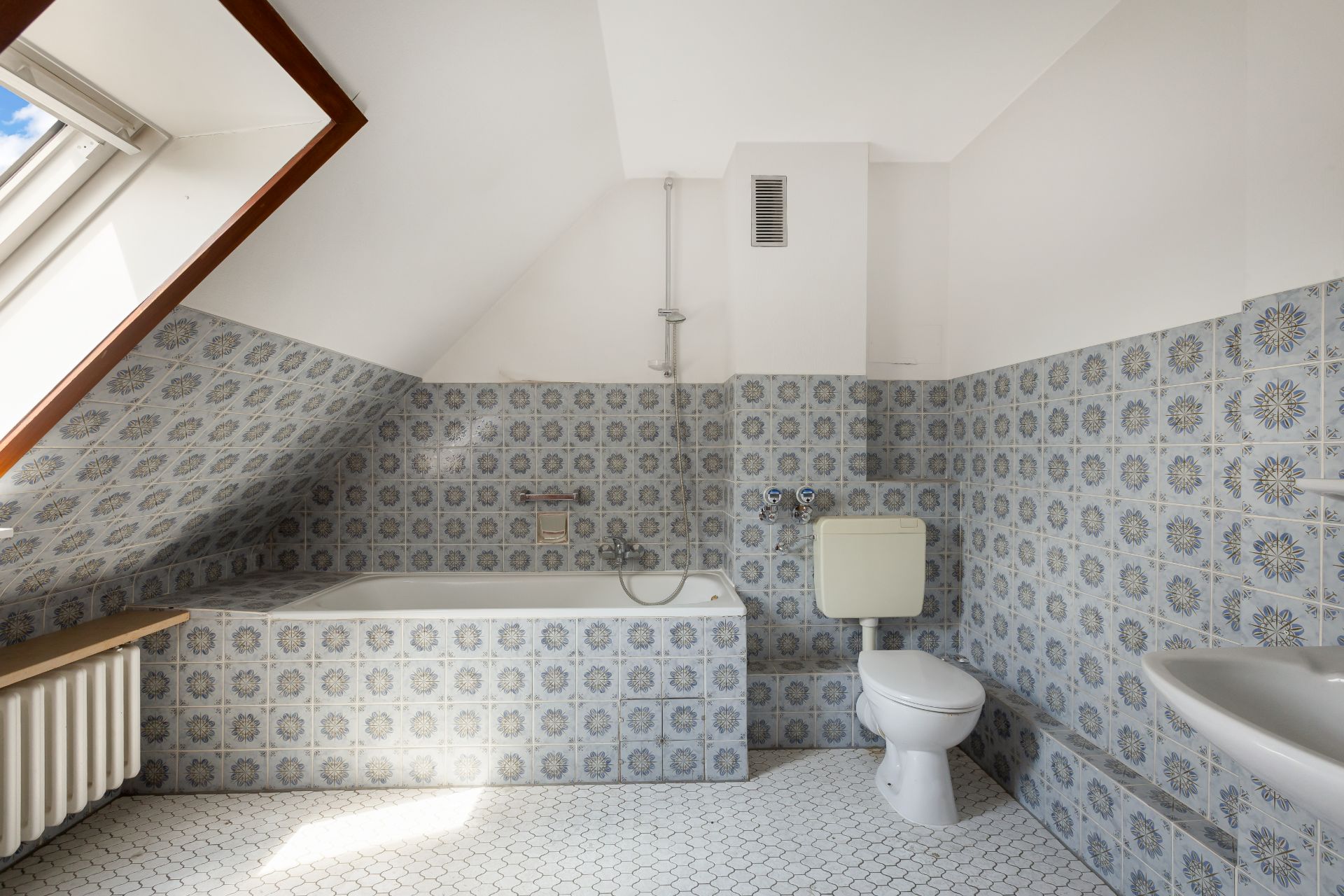 Badezimmer mit Badewanne, Toilette, Waschbecken und gemusterten Fliesen unter einer Dachschräge.