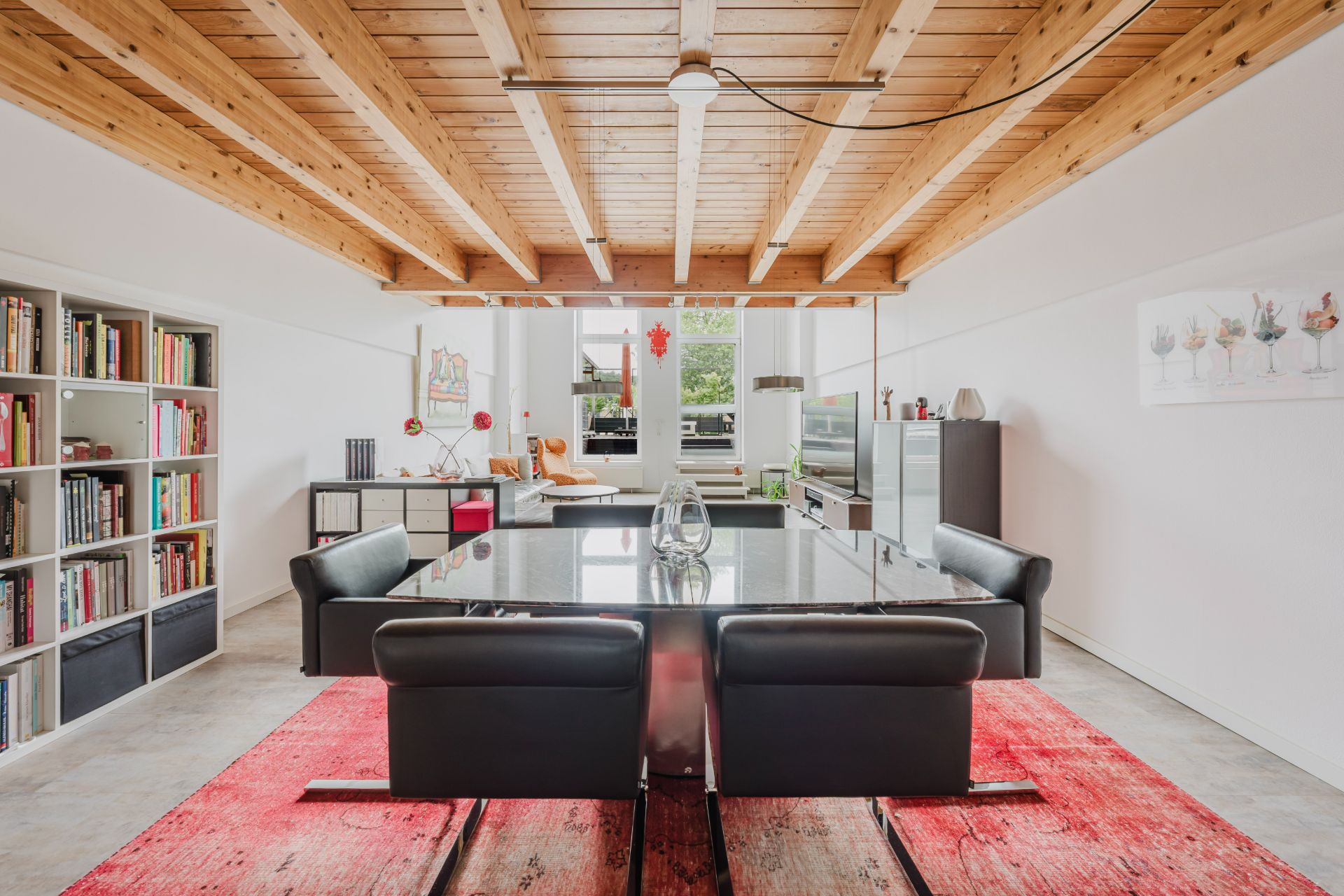 Helles Esszimmer mit Holzbalkendecke, großem Tisch, schwarzen Stühlen und roten Teppichen.