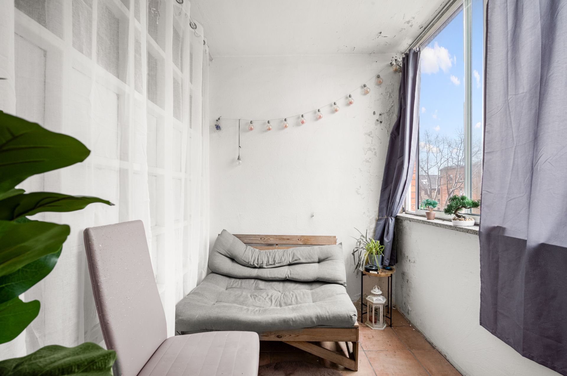 Kleiner Balkon mit Sitzbank, Stuhl, dekorativen Pflanzen und Blick durch ein Fenster.