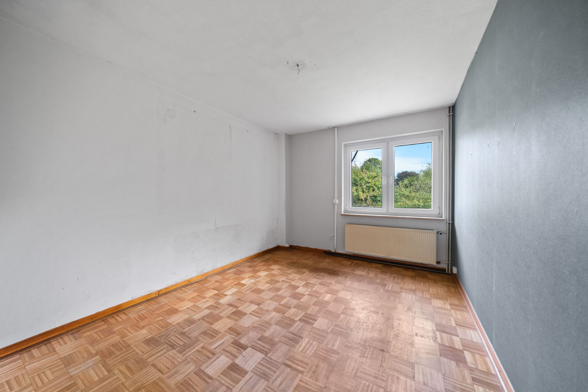 Leerer Raum mit Parkettboden, grauer Wand und einem Fenster mit Blick ins Grüne.