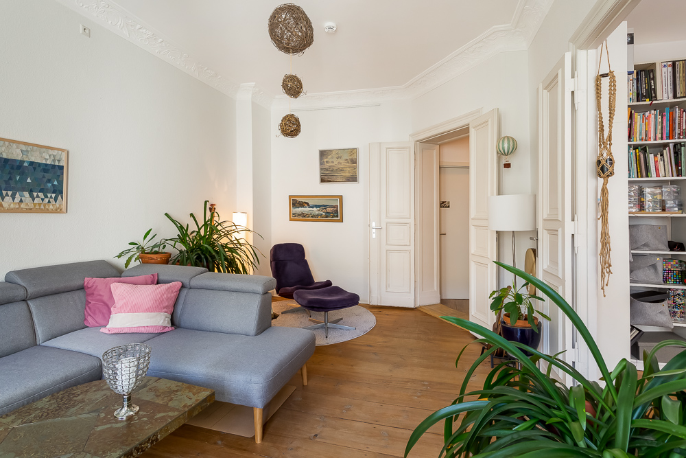 Helles Wohnzimmer mit grauem Sofa, Pflanzen, Bücherregal und Bildern an der Wand.