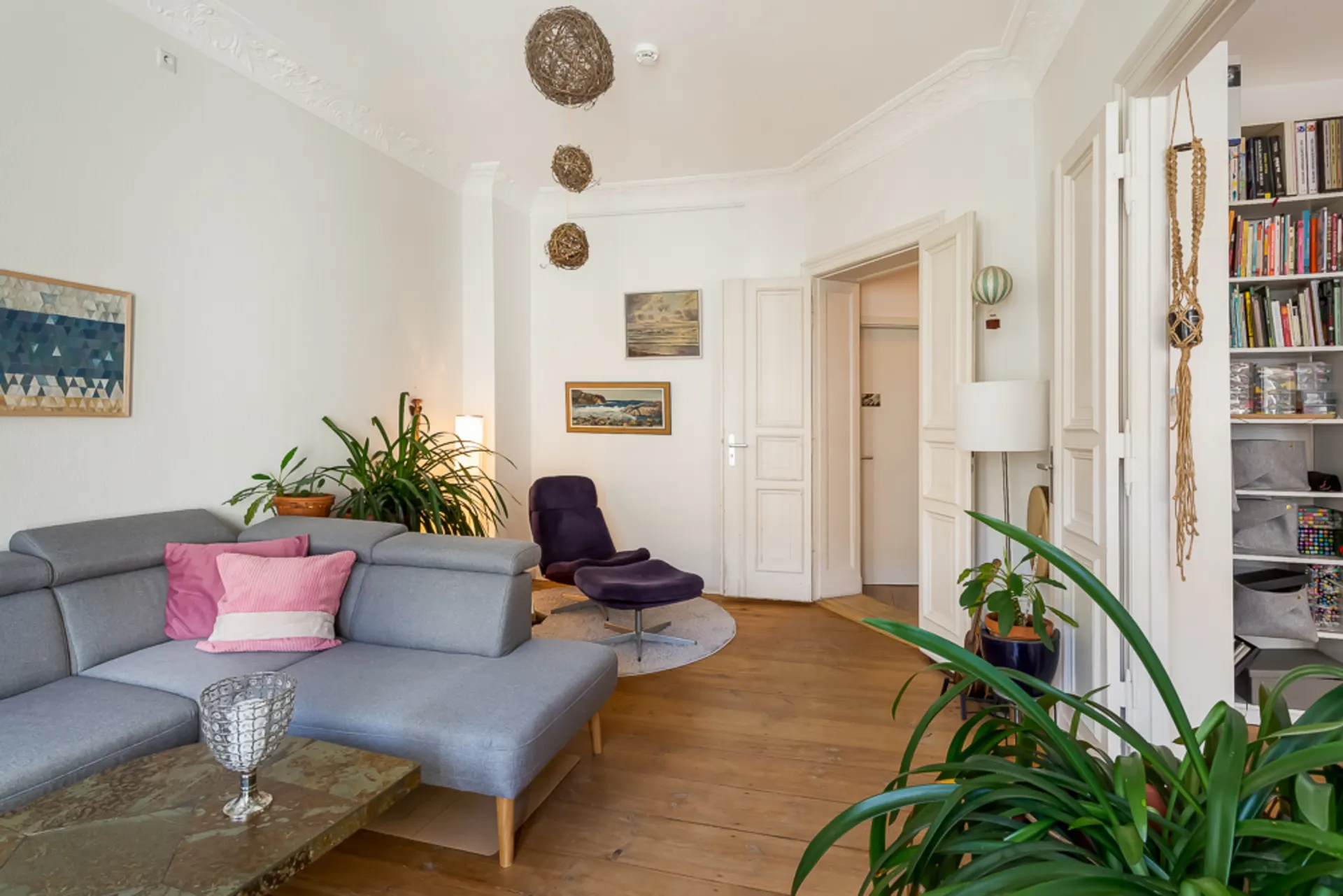 Helles Wohnzimmer mit grauem Sofa, Pflanzen, Bücherregal und Bildern an der Wand.