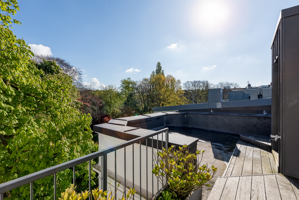 Kleine Dachterrasse mit Geländer, Holzboden und Pflanzen, Blick auf Bäume und Dächer bei Sonnenschein.
