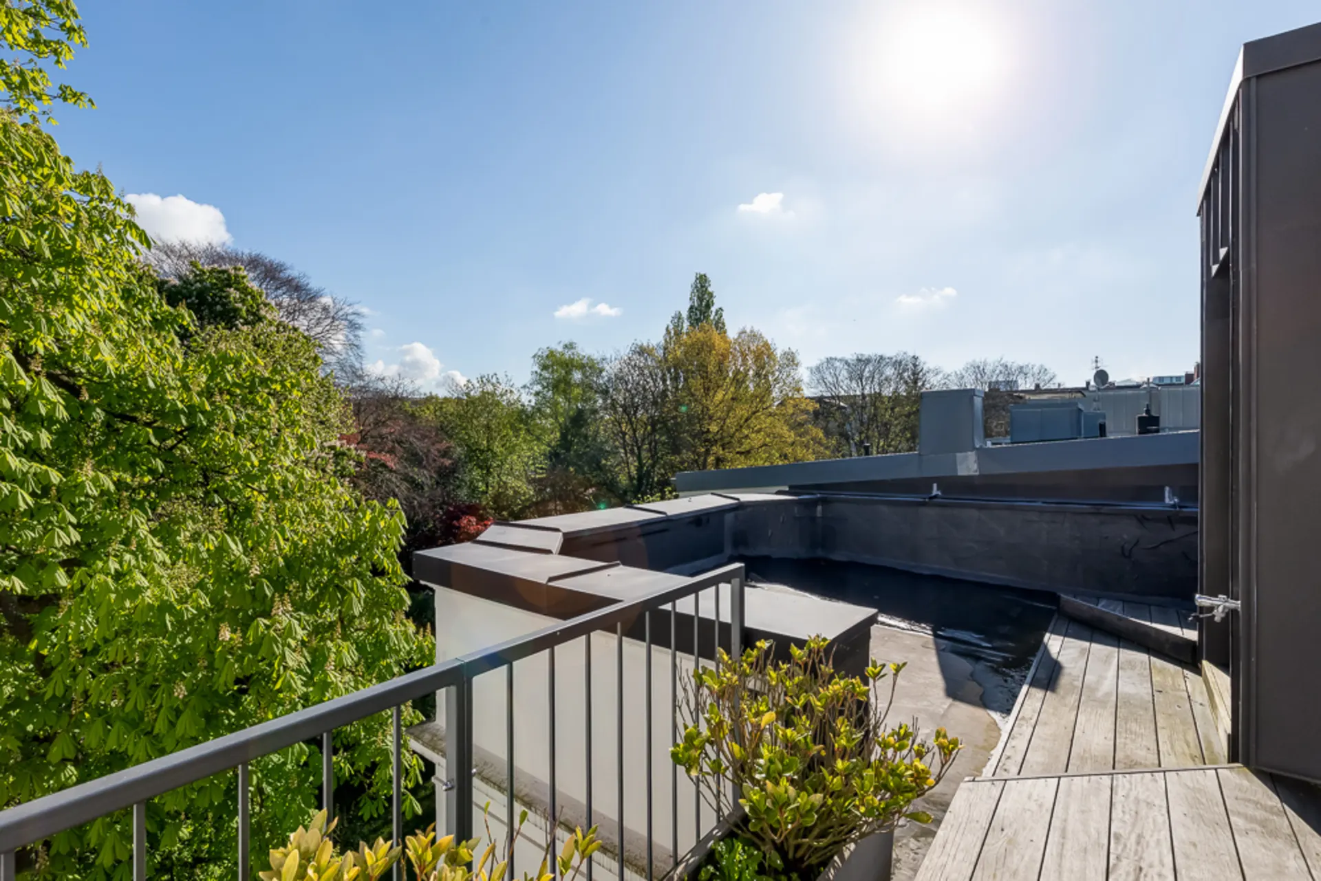 Kleine Dachterrasse mit Geländer, Holzboden und Pflanzen, Blick auf Bäume und Dächer bei Sonnenschein.