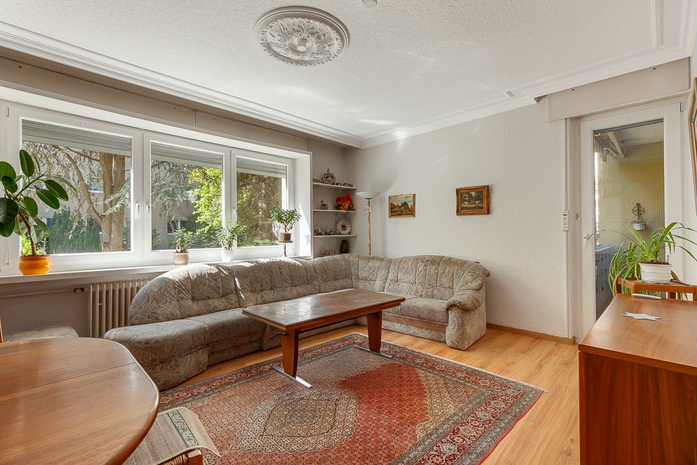 Helles Wohnzimmer mit Ecksofa, Holztisch, Teppich und großen Fenstern, Pflanzen auf der Fensterbank.
