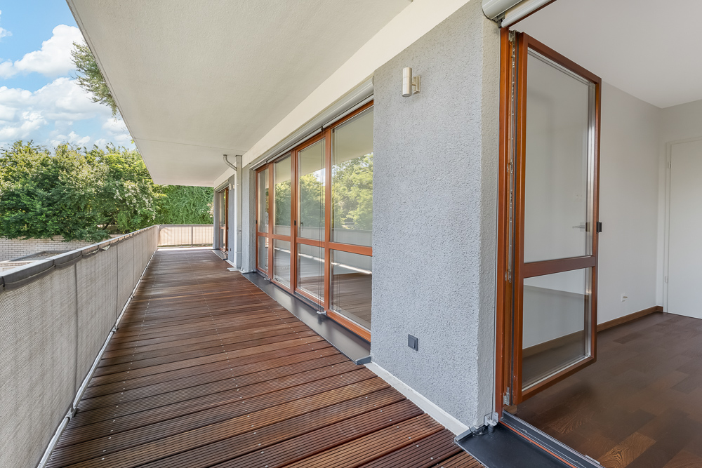 Großer Balkon mit Holzboden und Fensterfront, daneben eine geöffnete Tür ins Innere einer Wohnung.