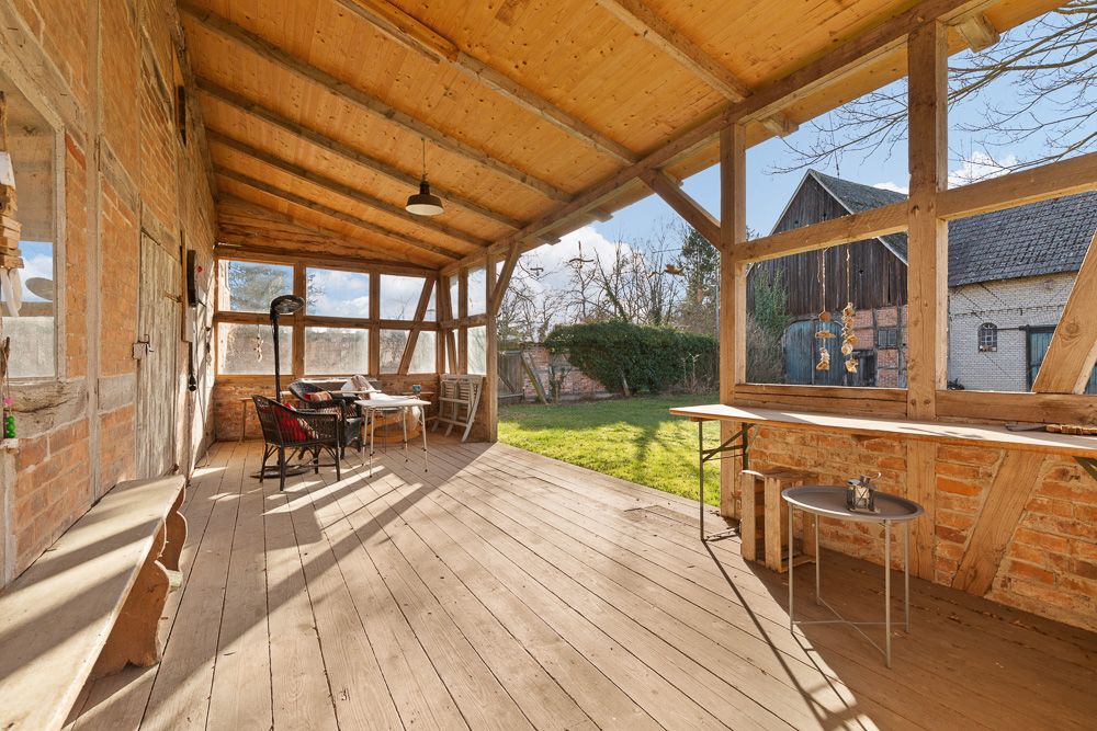 Überdachte Terrasse mit Holzboden, Sitzgruppe und Blick in einen Garten mit Scheunen im Hintergrund.