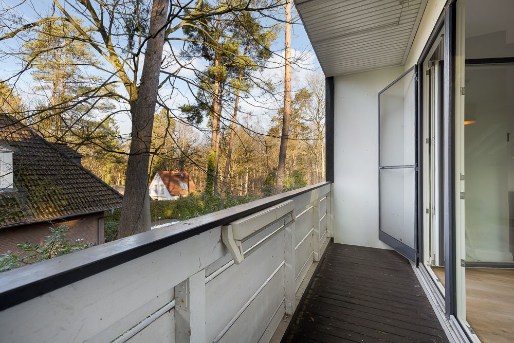 Balkon mit Holzboden, weißem Geländer und Blick auf Bäume, daneben eine geöffnete Glastür.