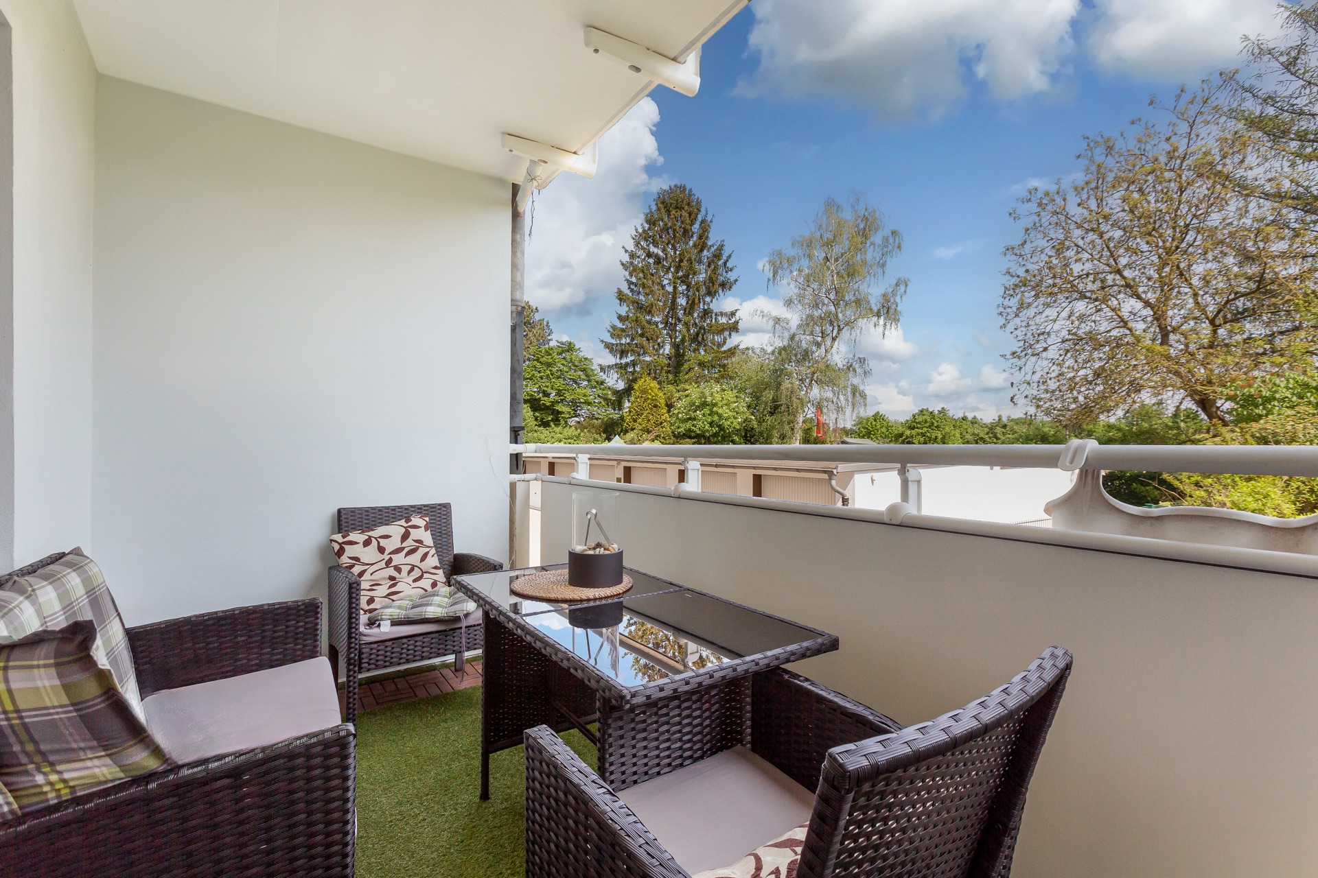 Balkon mit Rattanmöbeln, Glastisch und Blick auf Bäume bei blauem Himmel.