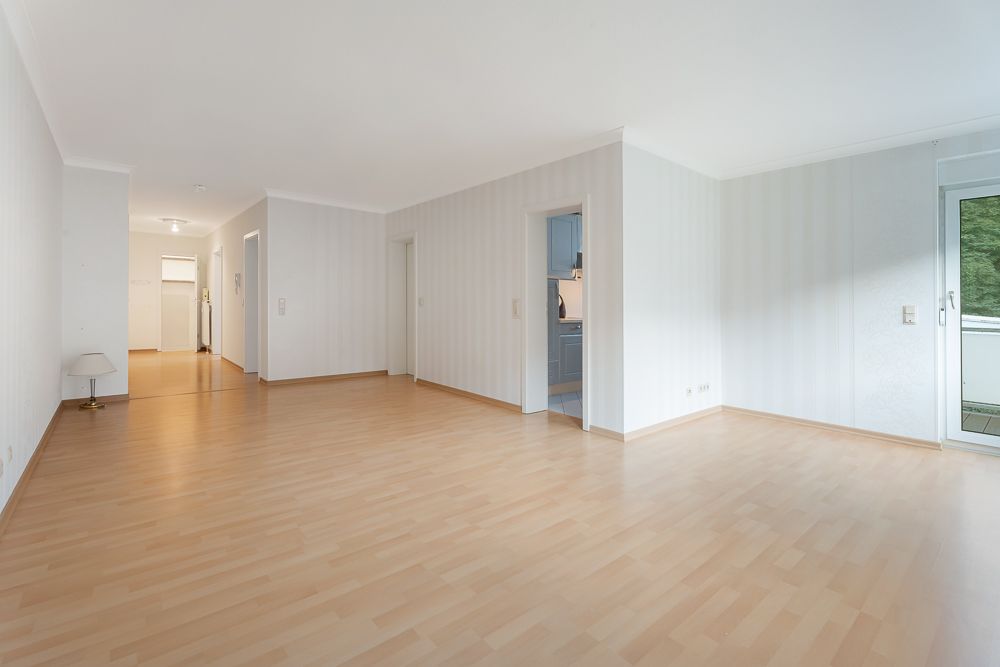 Helles, leeres Wohnzimmer mit Holzfußboden, zwei Türen und Zugang zu einem Balkon.