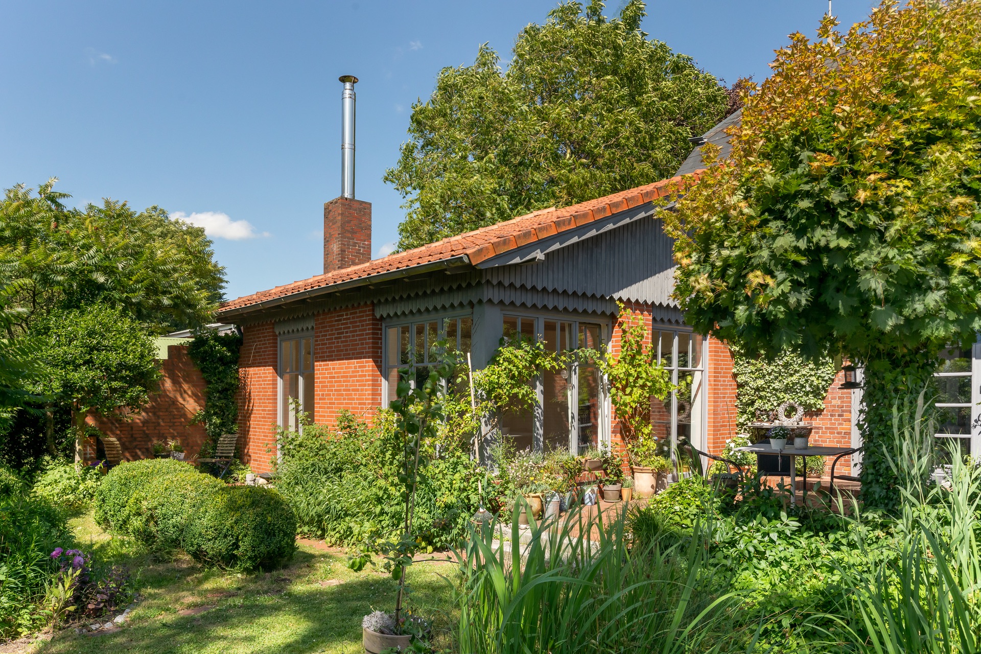Backsteinhaus mit Terrasse, Garten und vielen Pflanzen bei sonnigem Wetter.