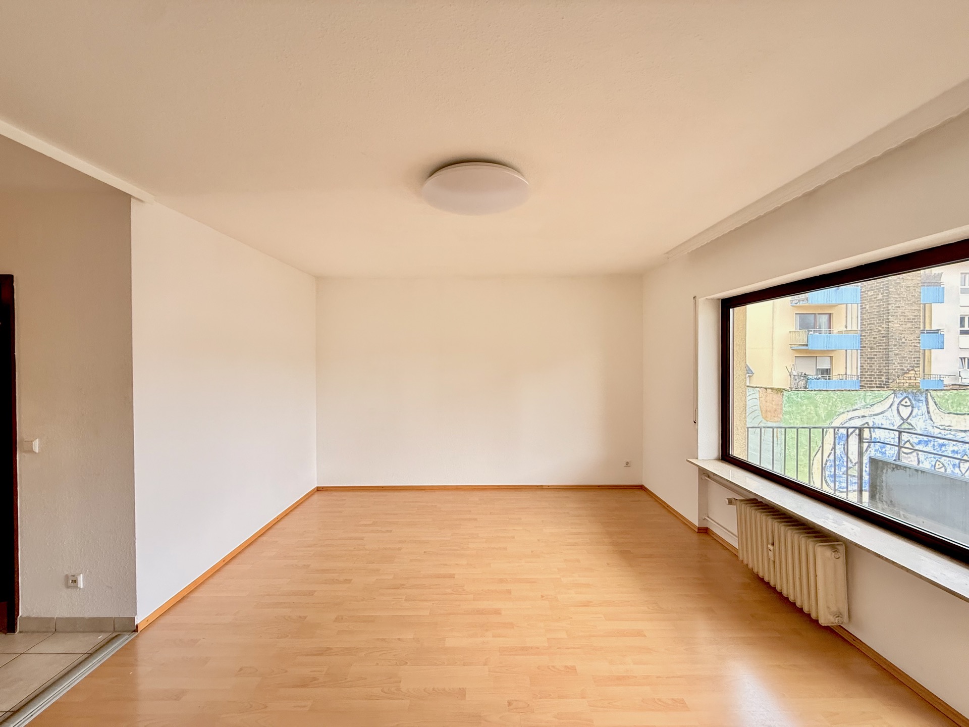 Helles, leeres Zimmer mit Holzfußboden und großem Fenster zur Straße.