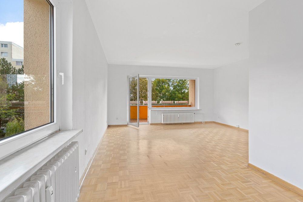 Helles, leeres Wohnzimmer mit Parkettboden und Zugang zu einem Balkon.