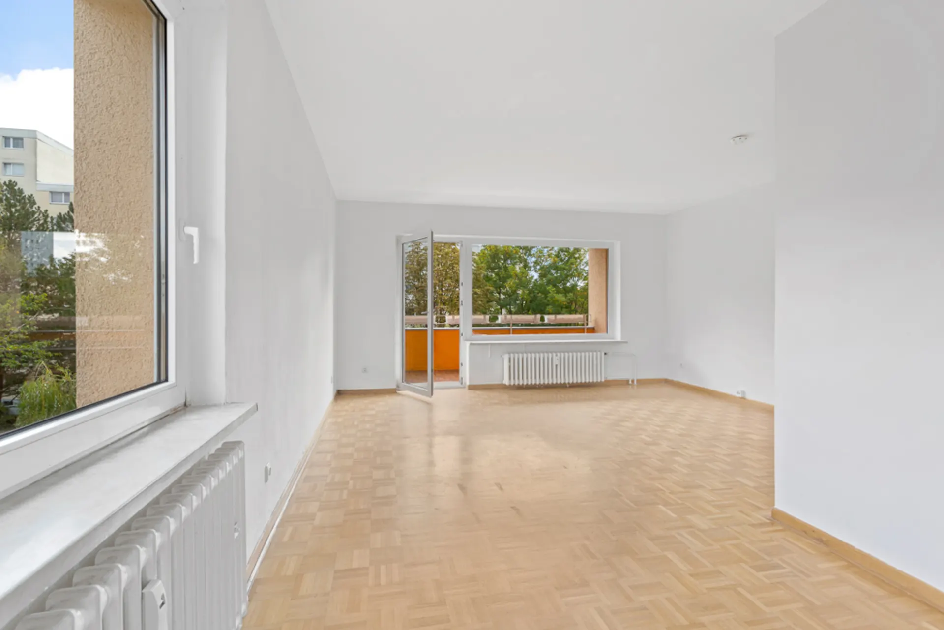 Helles, leeres Wohnzimmer mit Parkettboden und Zugang zu einem Balkon.