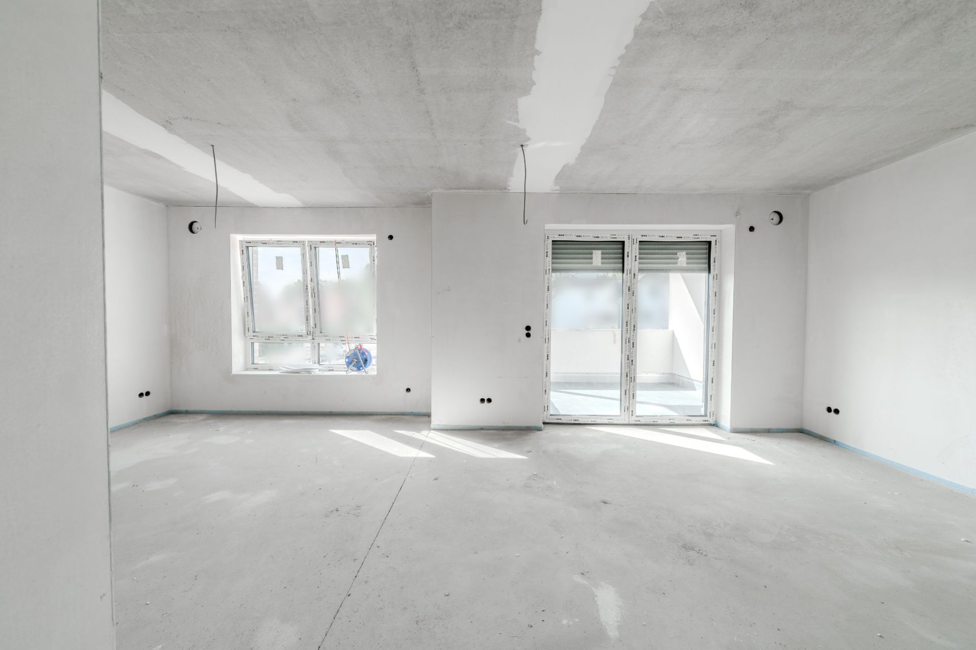 Leerer, weiß gestrichener Raum mit zwei Fenstern und Glastür zu einem Balkon.