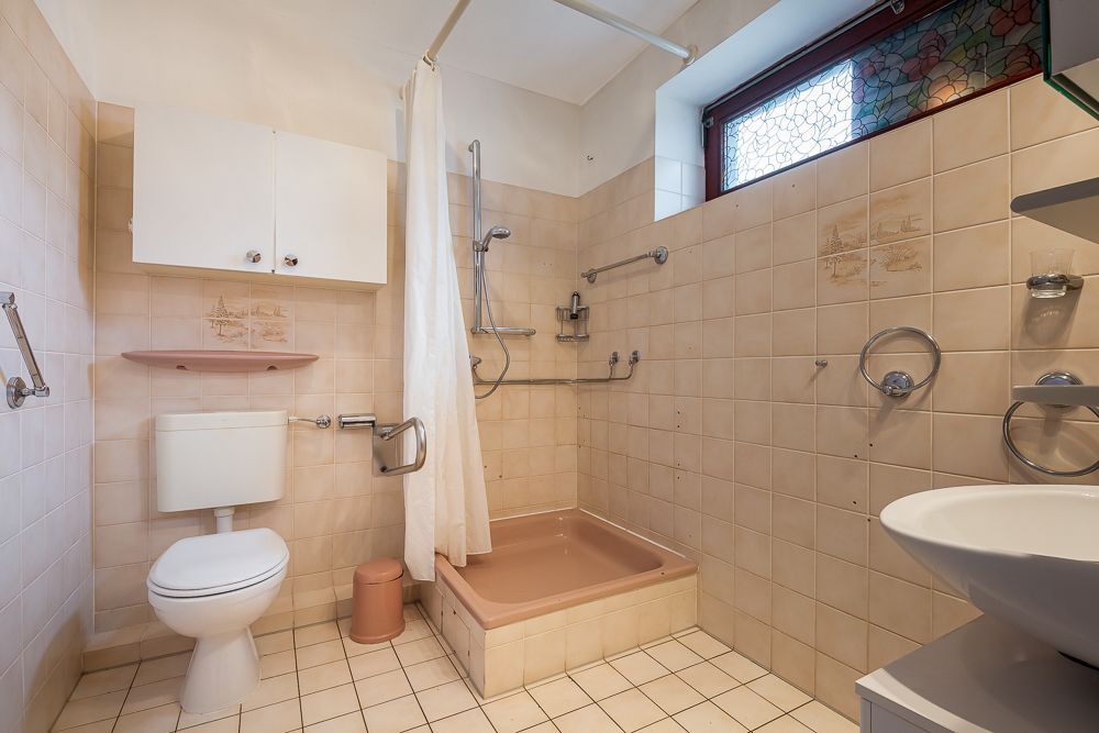 Badezimmer mit beiger Fliesen, begehbarer Dusche, WC, Waschbecken und Haltegriffen.