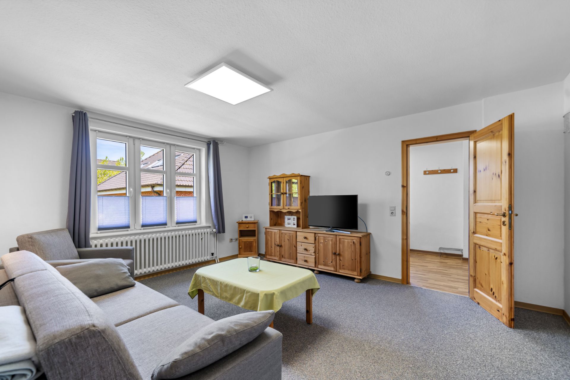 Wohnzimmer mit Sofa, Couchtisch, Fernseher auf Holzschrank und großem Fenster mit Vorhängen.