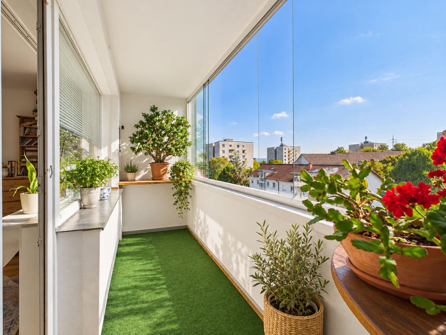 Heller, verglaster Balkon mit grünen Pflanzen, Kunstrasen und Blick auf Wohngebäude.