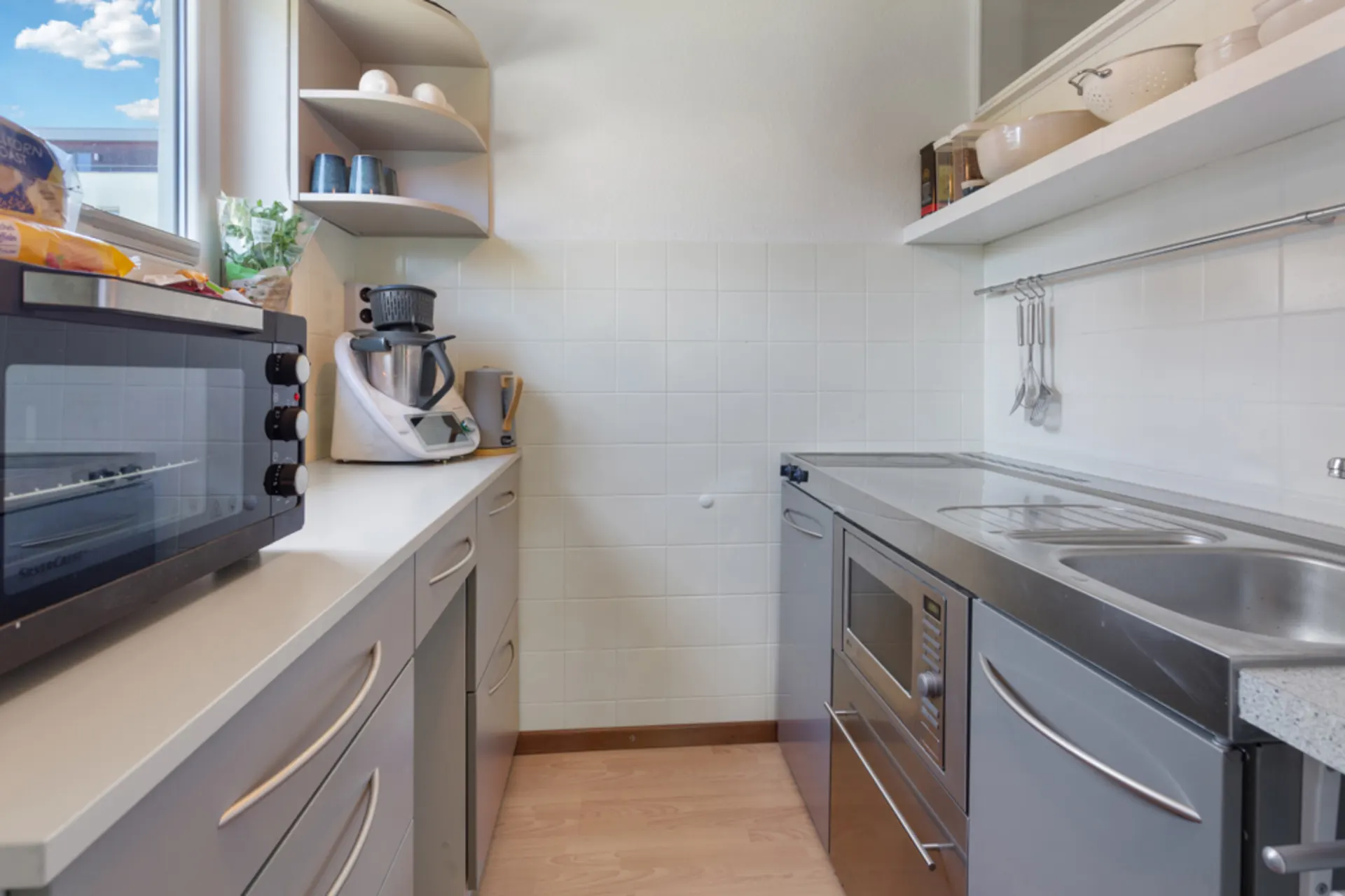 Moderne, schmale Küche mit Küchengeräten, grauen Schränken und einem Fenster.