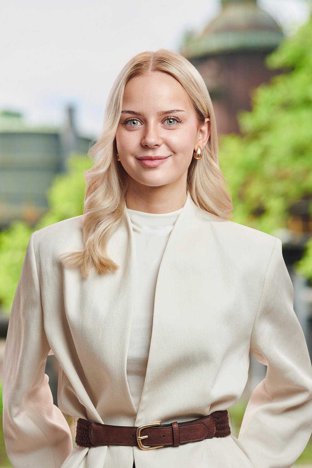 Junge Frau mit blondem Haar und weißem Blazer steht draußen vor grünen Bäumen.