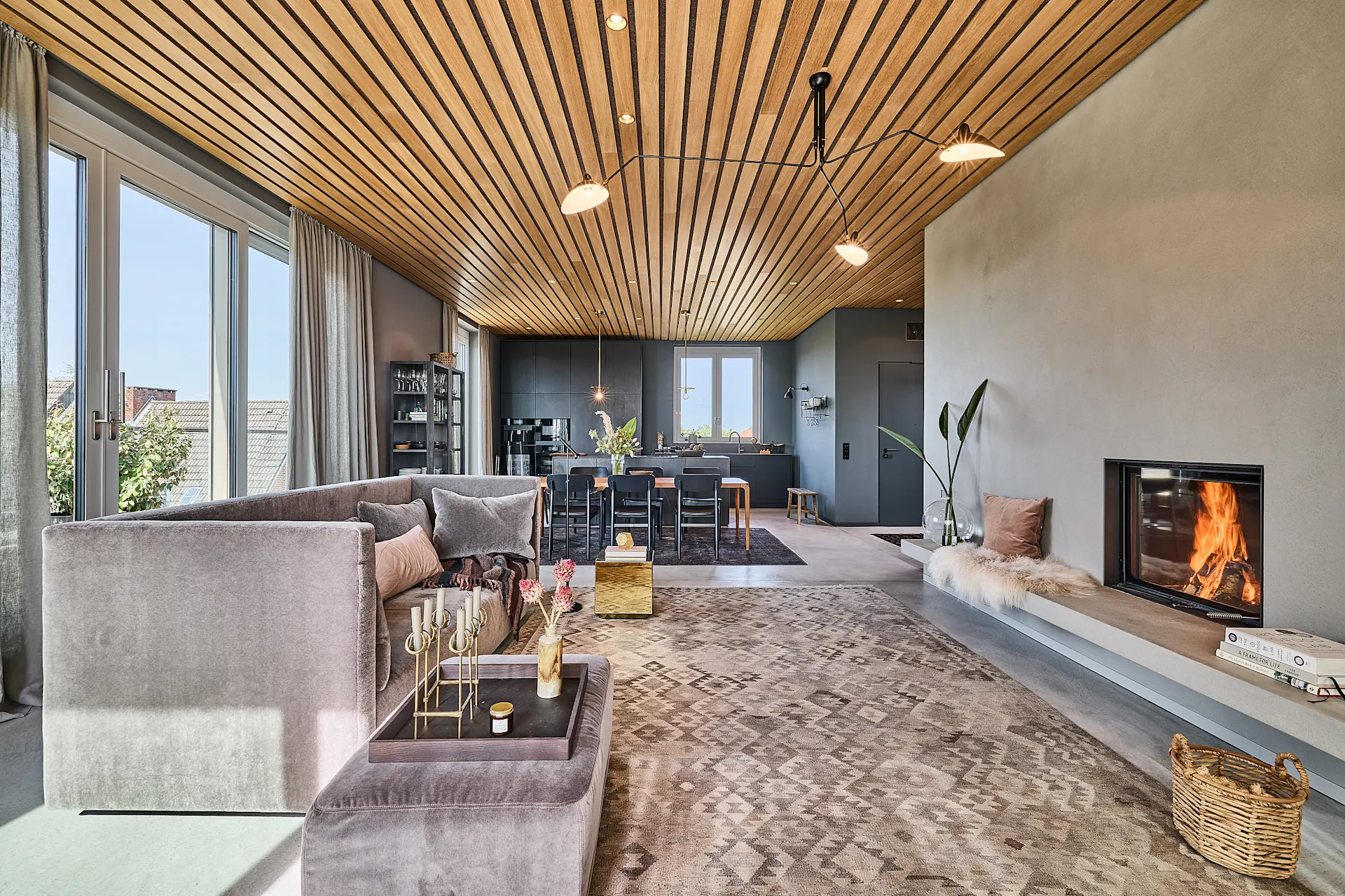Modernes Wohnzimmer mit Kamin, großem Sofa, Teppich und offener Küche im Hintergrund.