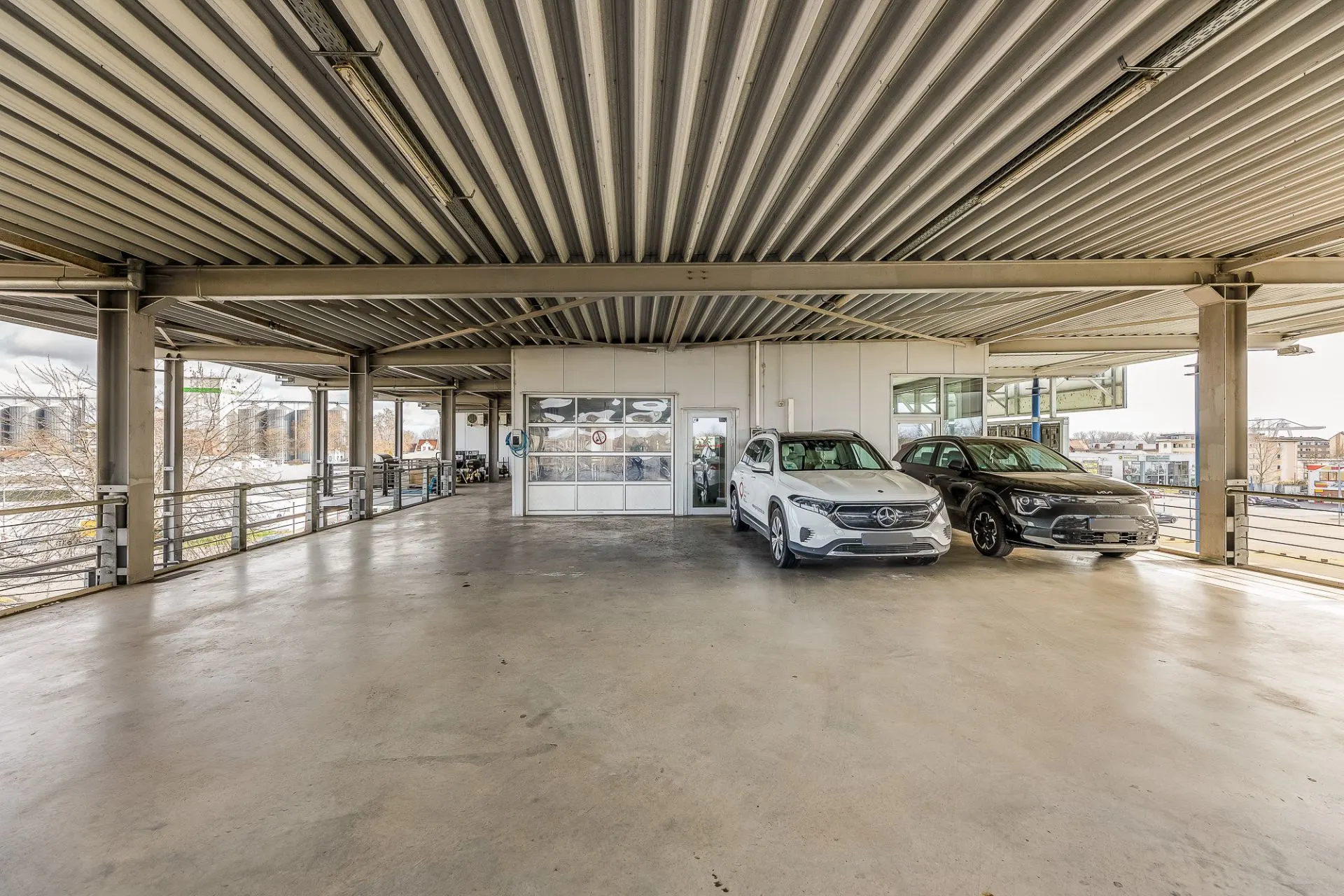 Überdachter Parkplatz mit zwei geparkten Autos und einem kleinen Gebäude im Hintergrund.