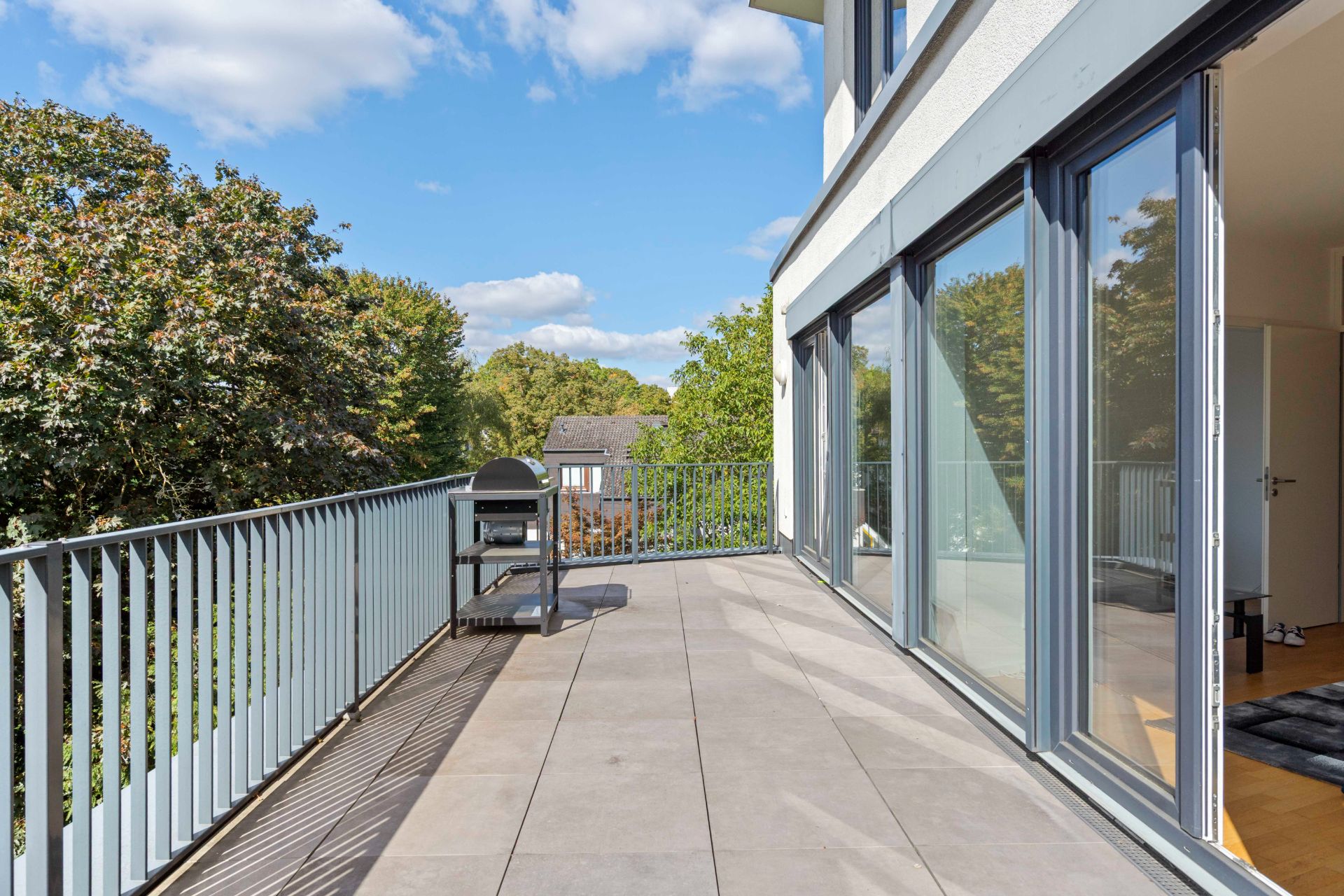 Großer Balkon mit Fliesen, Geländer, Grill und bodentiefen Fenstern, mit Blick auf Bäume.