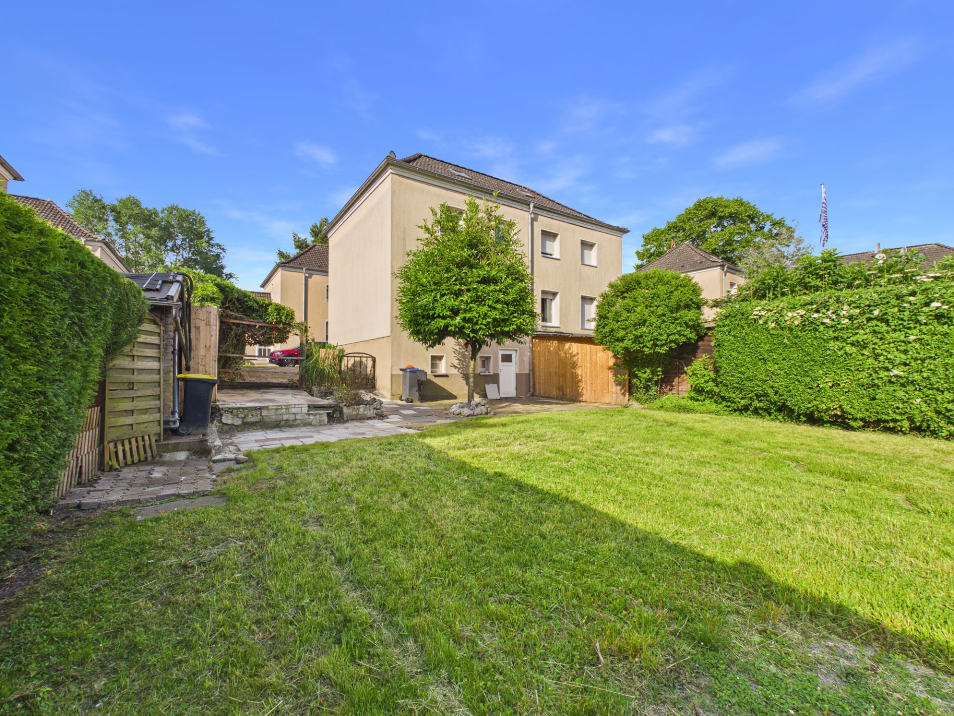 Garten mit Rasenfläche, zwei Bäumen und einem Mehrfamilienhaus im Hintergrund.