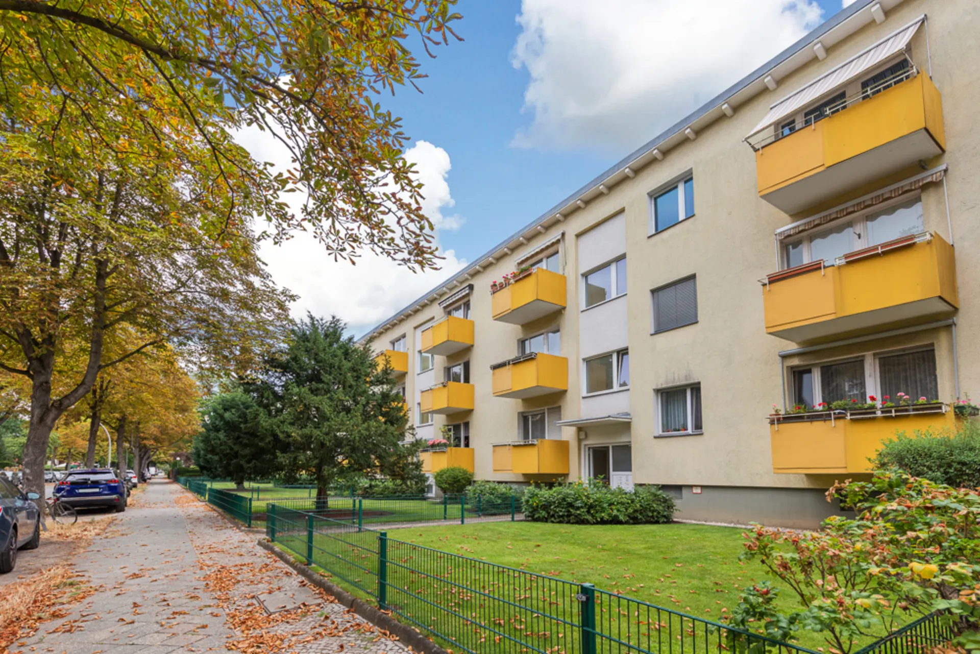 Mehrfamilienhaus mit gelben Balkonen, Bäumen und Gehweg im Vordergrund.