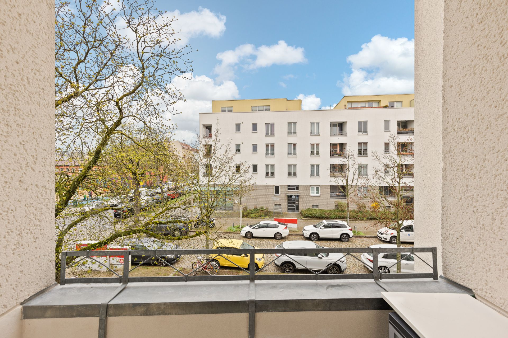 Blick von einem Balkon auf eine Straße mit geparkten Autos und einem Wohnhaus gegenüber.