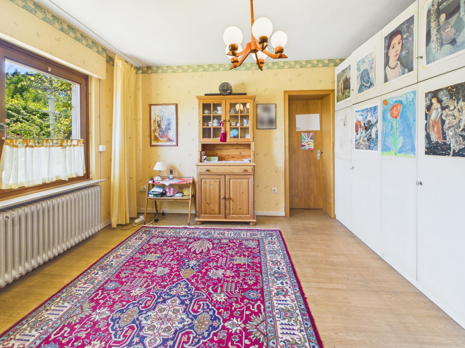 Helles Zimmer mit großem Fenster, Teppich, Holzschrank, Malereien an weißen Schränken und Deckenleuchte.