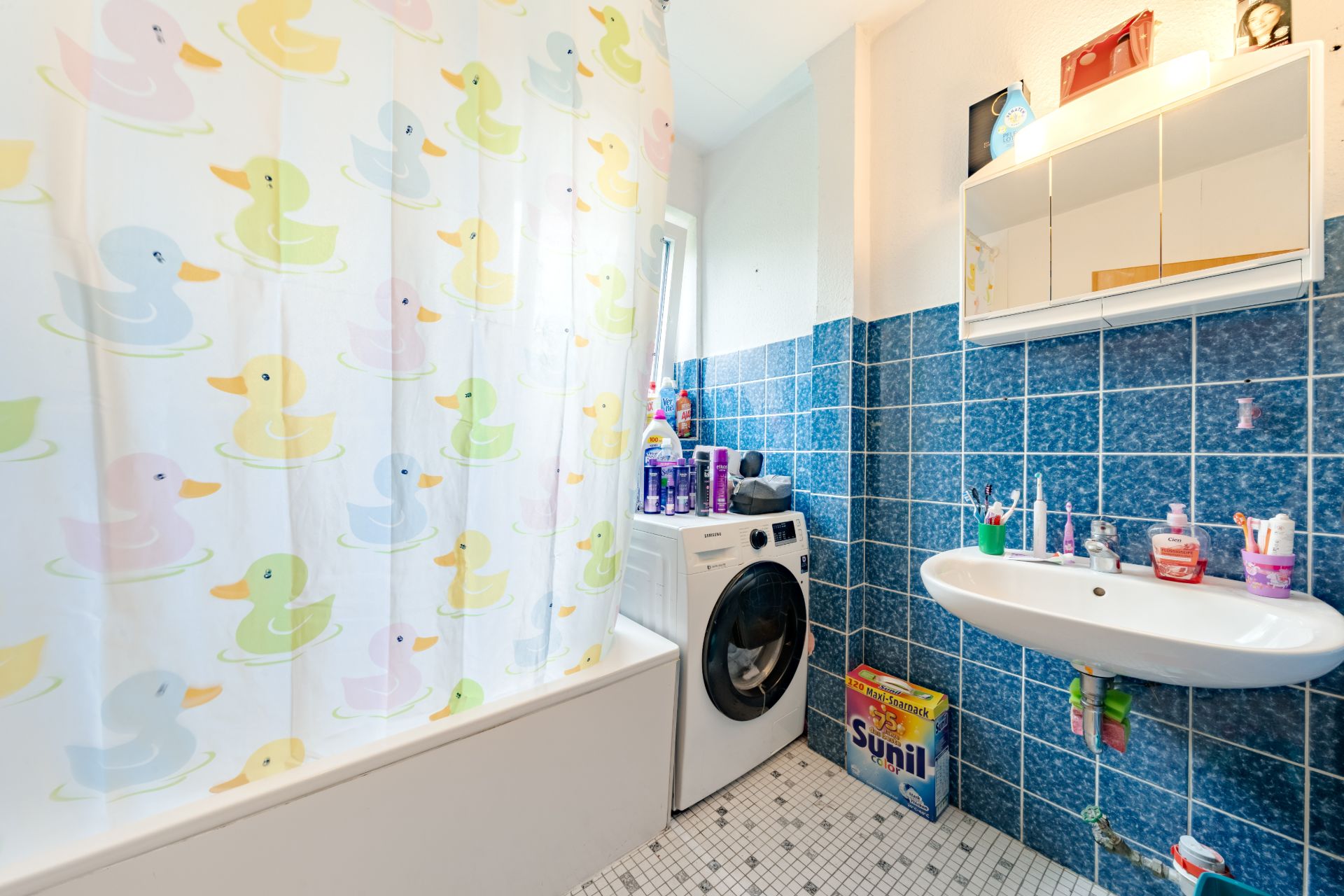 Badezimmer mit blau gefliester Wand, Waschmaschine, Waschbecken und bunter Duschvorhang mit Enten.