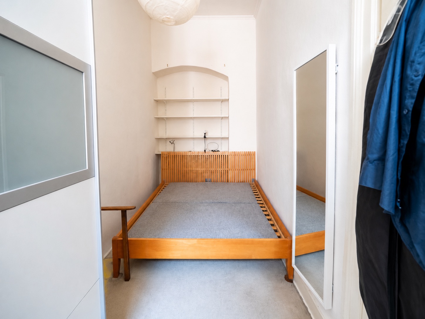 Schmales Schlafzimmer mit Holzbettgestell, Regal an der Wand und großem Spiegel.