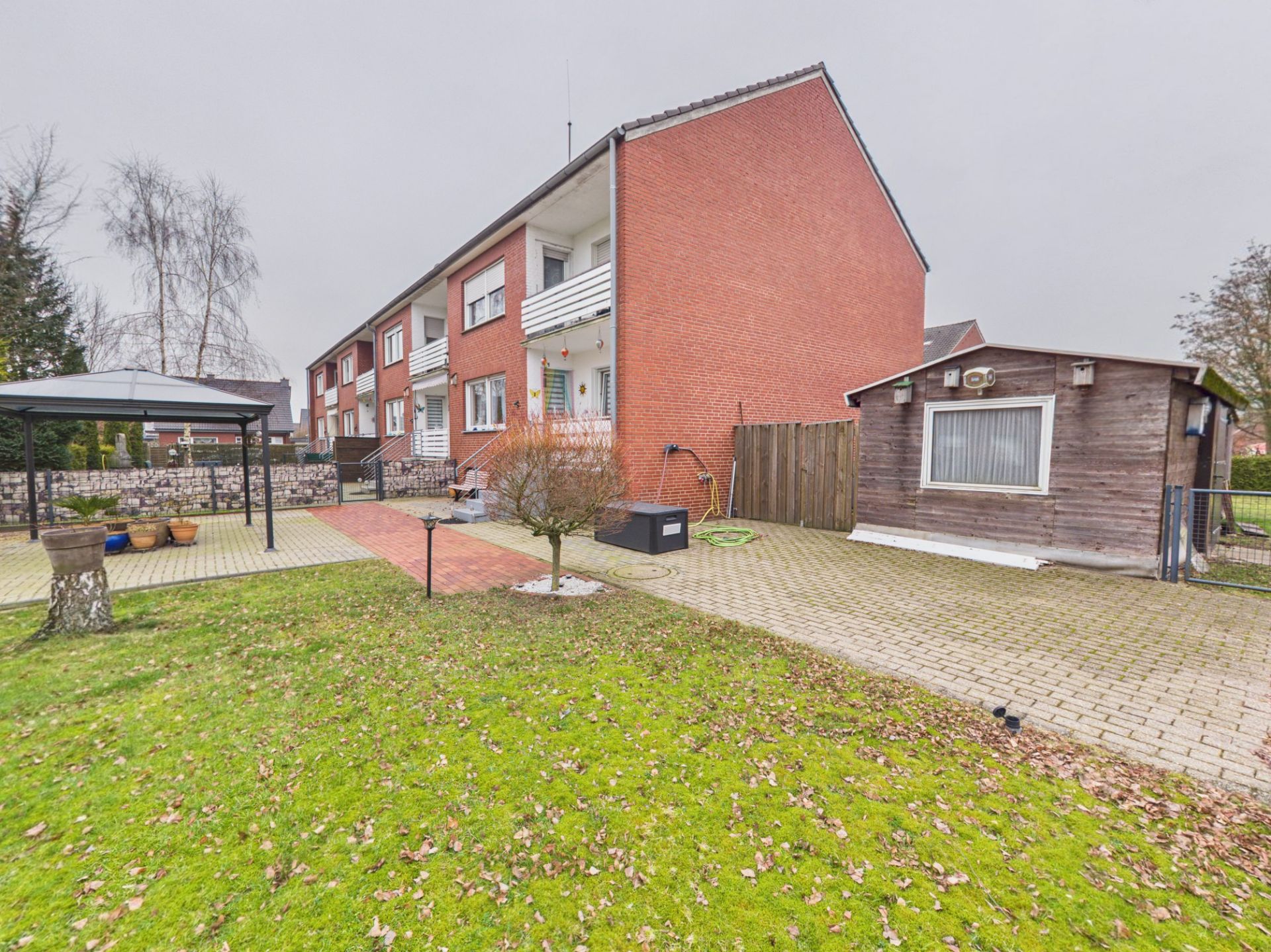 Garten mit Rasenfläche, Pavillon, Nebengebäude und rotem Mehrfamilienhaus im Hintergrund.