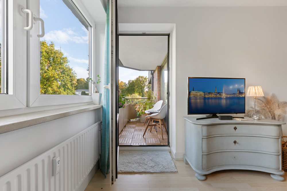 Wohnzimmer mit offenem Fenster, Blick auf Balkon mit Stuhl und Pflanze, Fernseher auf Kommode.
