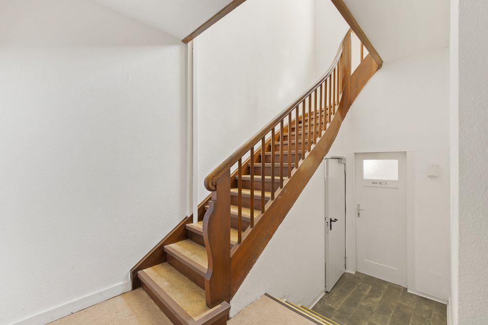 Holztreppe in einem hellen Flur mit weißer Tür unten rechts.