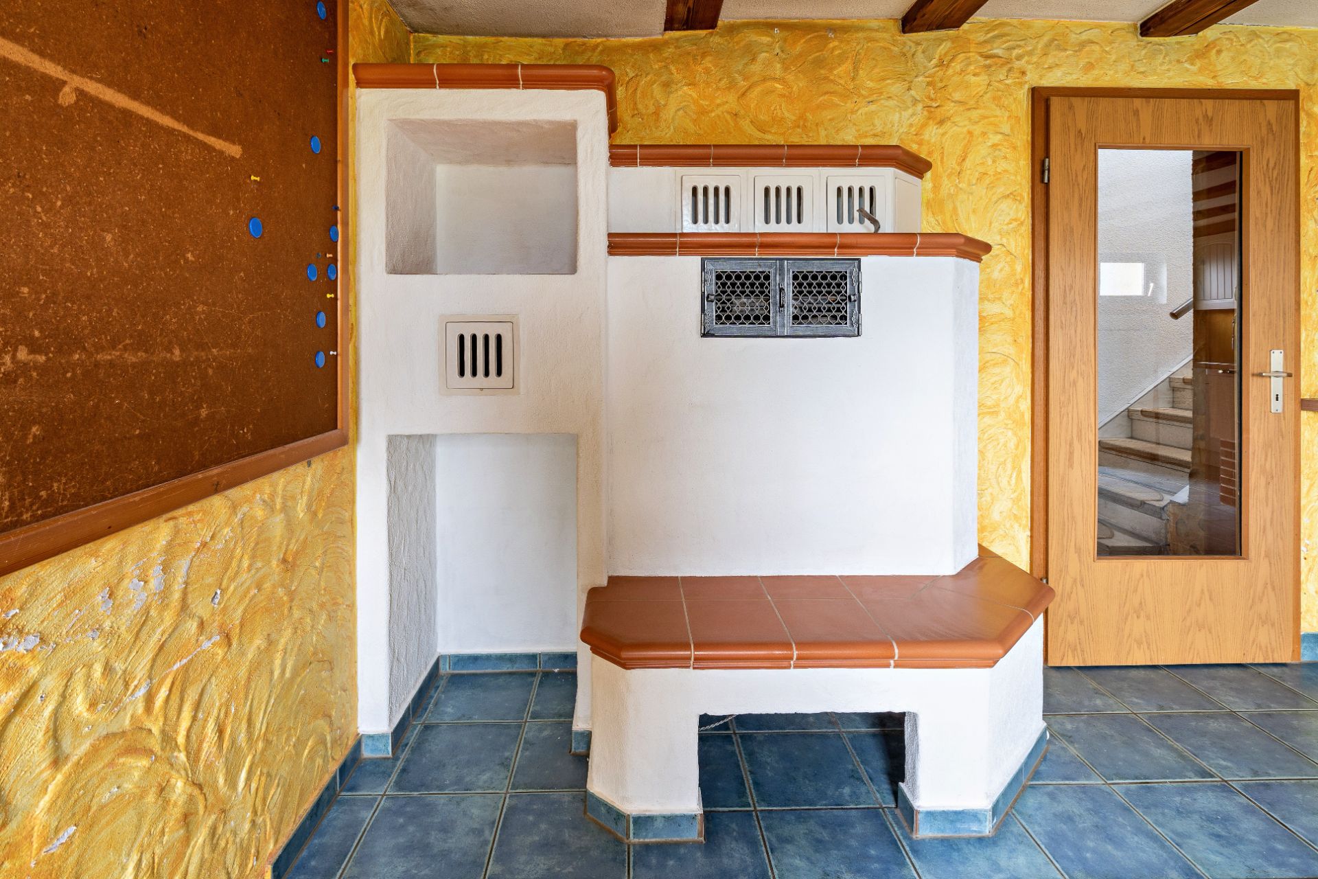 Innenraum mit gemauertem Sitzplatz, gelber Wand, Korkpinnwand und Holztür mit Glasfenster.