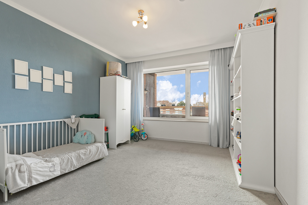 Helles Kinderzimmer mit Bett, Schrank, Regal und großem Fenster, Spielzeug auf dem Regal.