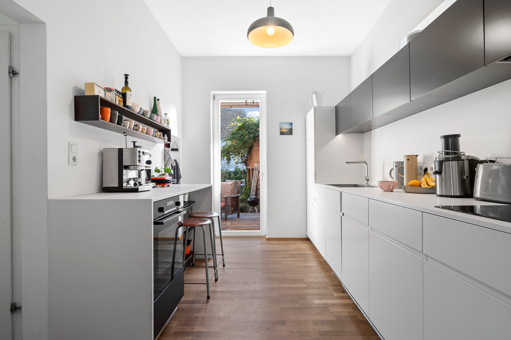 Moderne, schmale Küche mit weißen Schränken, offenen Regalen und Blick auf eine Terrasse.
