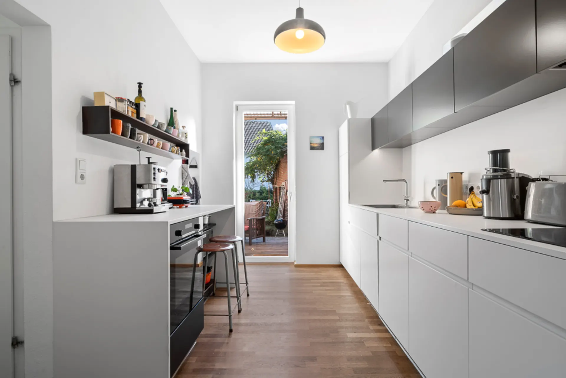 Moderne, schmale Küche mit weißen Schränken, offenen Regalen und Blick auf eine Terrasse.